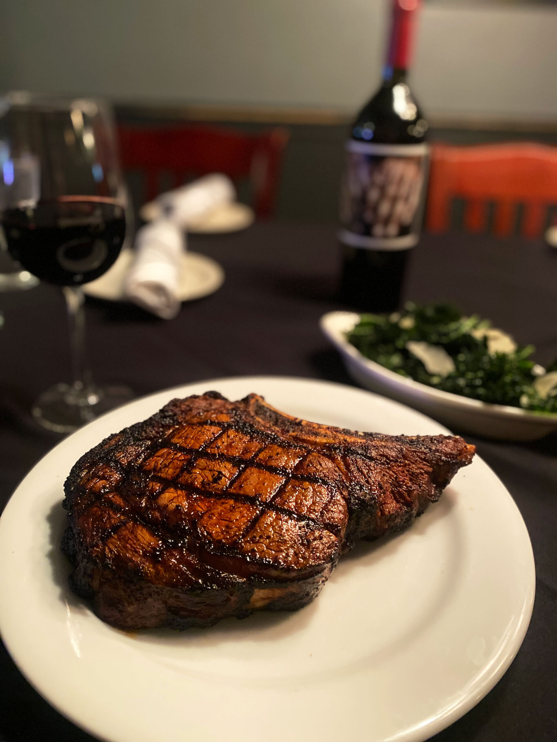 A large bone-in steak and red wine sit on a table with a black tablecloth.