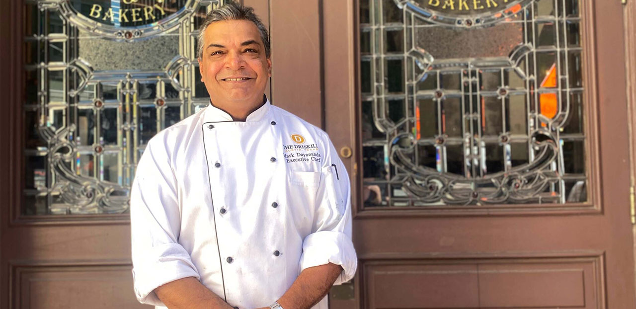 Executive Chef Mark Dayanandan standing outside of the Driskill Hotel in Austin, Texas smiling.